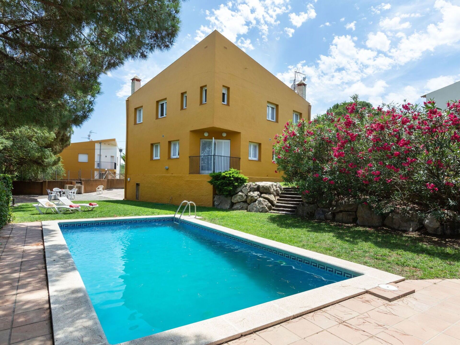 A yellow building with a swimming pool, surrounded by greenery and a pink flowering bush.