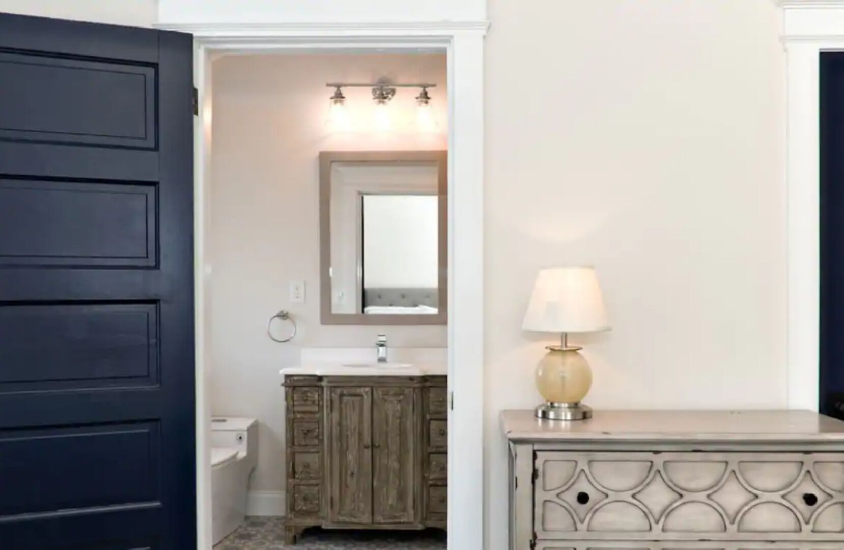 A bathroom with a dark blue door, a wooden vanity with a mirror, a white sink, and a beige lamp.