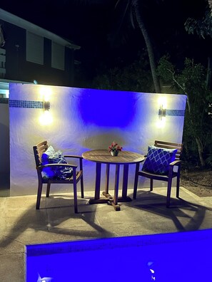 An outdoor seating area with a round table and two chairs, a small table with a flower vase, and a wall with two light fixtures. - House 2 min walk to Grace Bay Beach - 2b/1b, for 4, pool, AC, WiFi (The Bight Settlement)