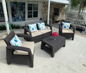 Un patio con mobiliario de mimbre, incluyendo dos sillones y un sofá, cada uno con cojines estampados en azul y blanco. En el centro hay una mesa pequeña con un jarrón de flores.