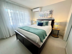A hotel room with a bed, a painting, an air conditioning unit, and a nightstand with a lamp.
