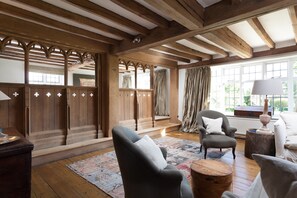 A room with exposed wooden beams, a large wooden cabinet with glass doors, a grey armchair with a white pillow, and a patterned rug on the wooden floor.
