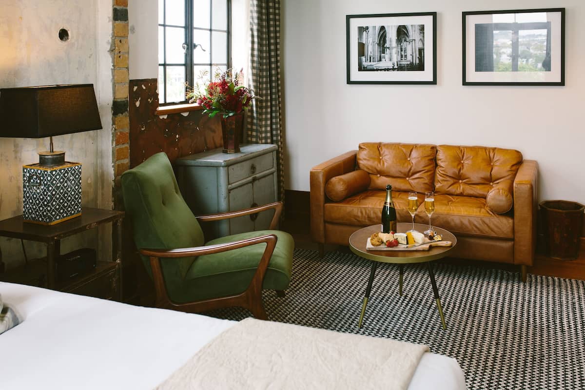 A hotel room with a bed, a green armchair, a brown leather sofa, a small table with a bottle and glasses, and two framed pictures on the wall.