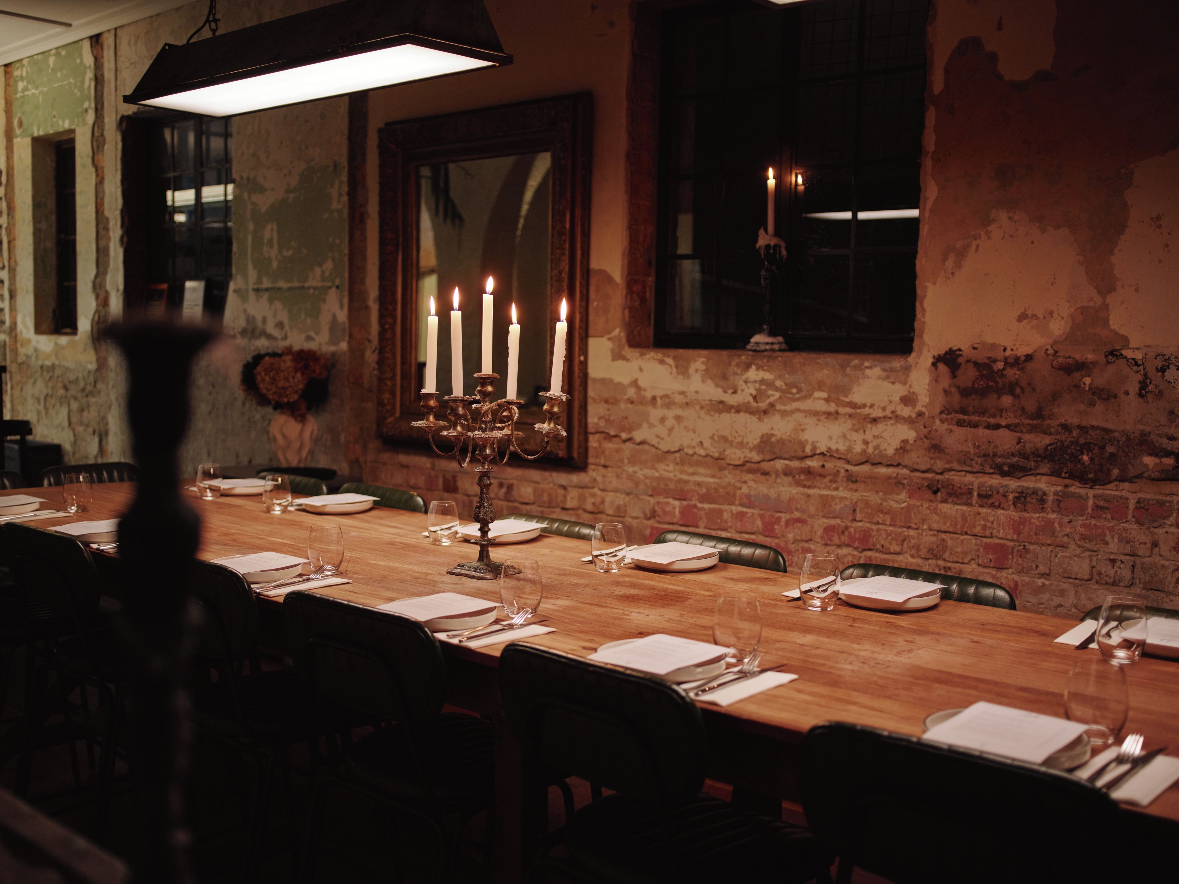 A long dining table set for a meal with candles, in a room with exposed brick walls and a large mirror.