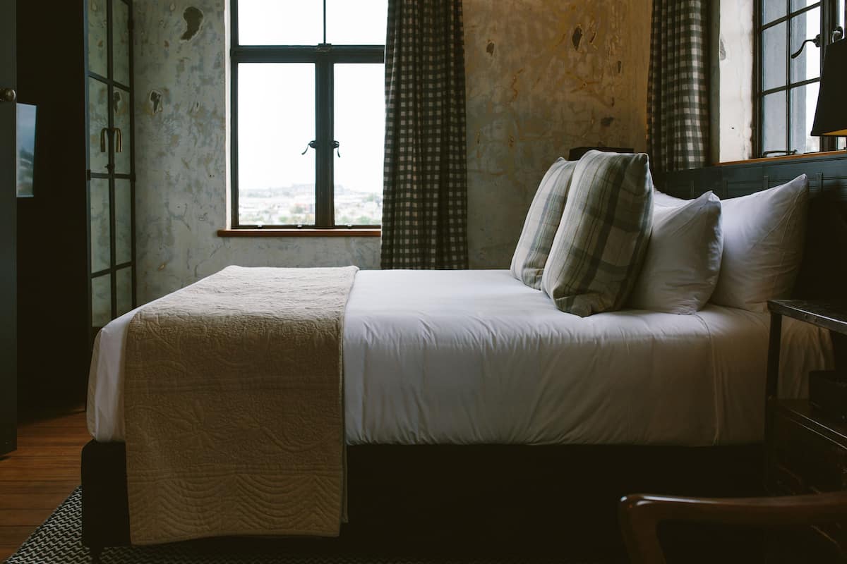 A bed with a quilt, a window with curtains, and a view of the outside.