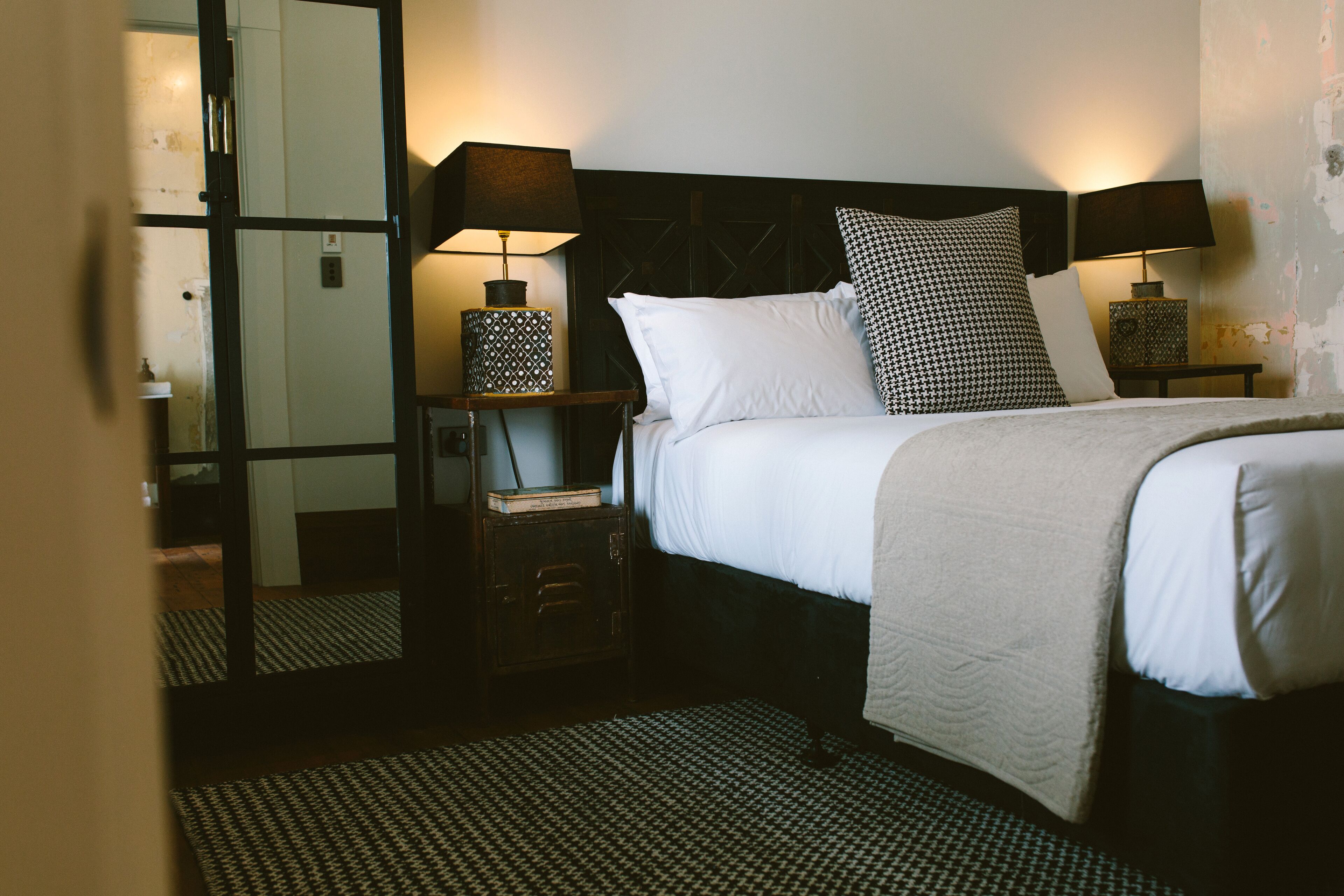 Habitación de hotel con una cama, dos mesitas de noche con lámparas, un espejo y un tapete estampado.