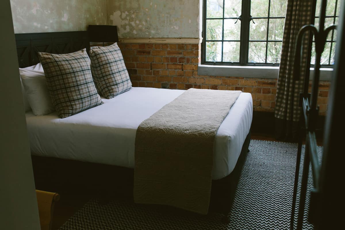A bed with a white sheet and two plaid pillows, a quilt, and a window with a view of trees.