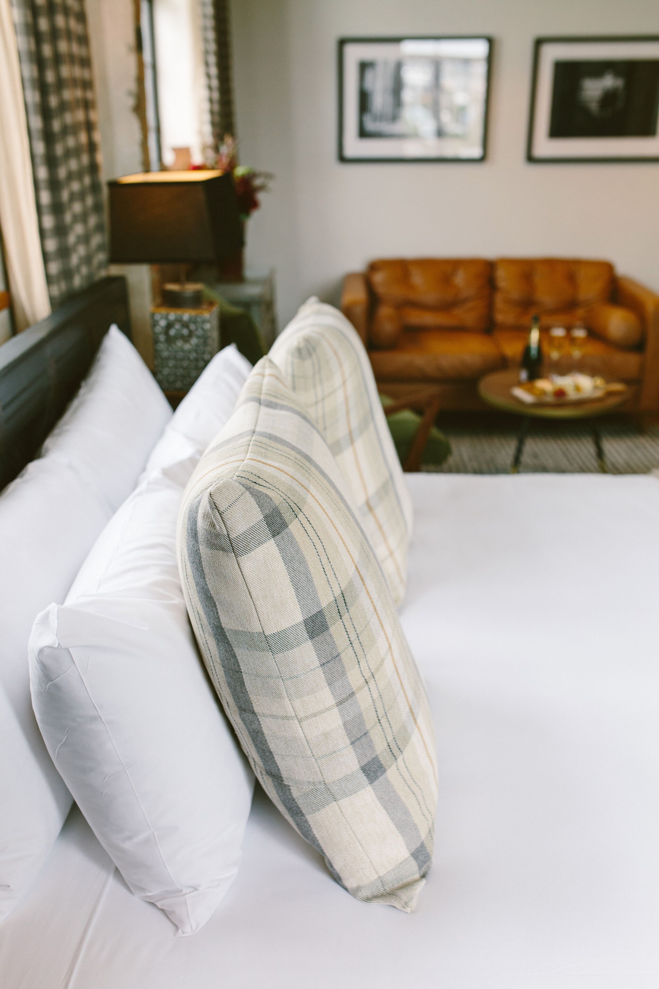 A neatly made bed with white and plaid pillows, a brown leather sofa, and a small round table with a bottle and glasses.