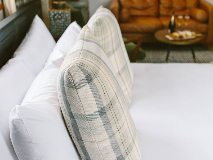 Una cama bien tendida con almohadas blancas y de cuadros, un sofá de cuero marrón y una mesita redonda con una botella y vasos.
