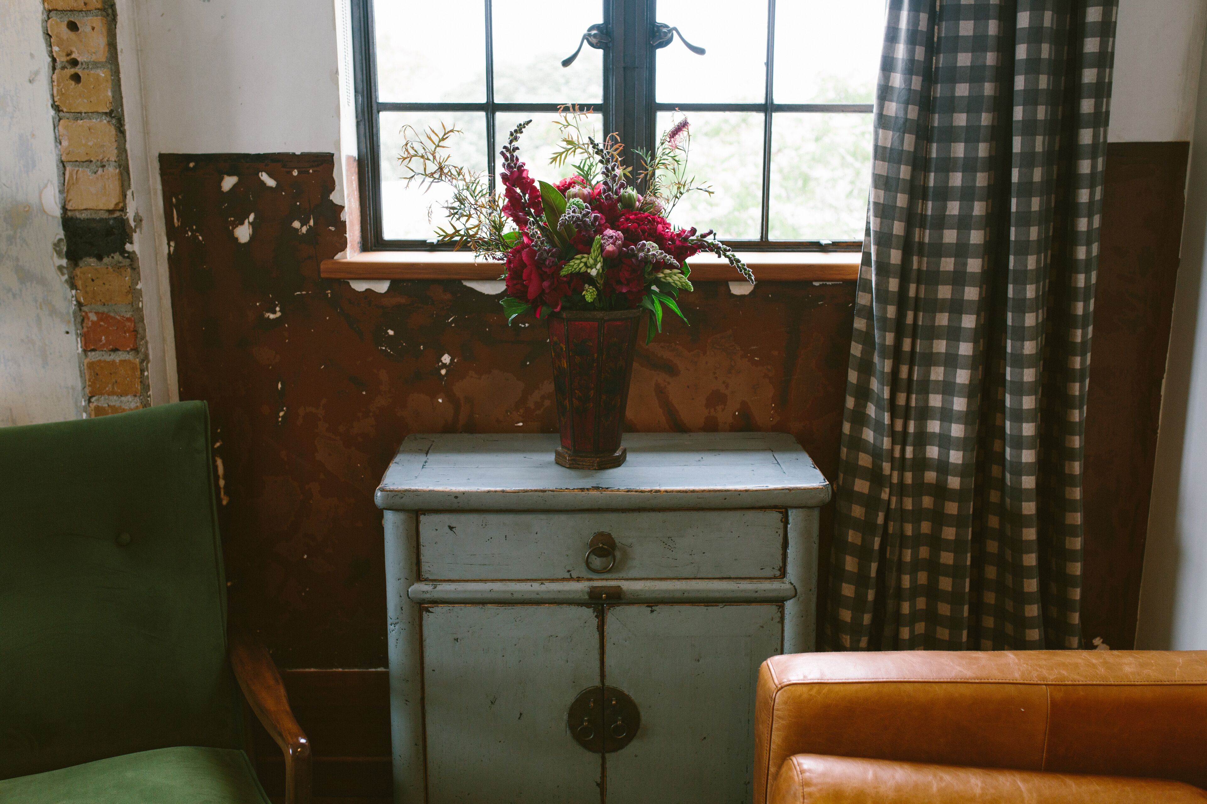 Ein antiker Schminktisch mit Blumenarrangements, ein grüner Sessel und eine braune Couch in einem Zimmer mit einem Fenster und karierten Vorhängen.