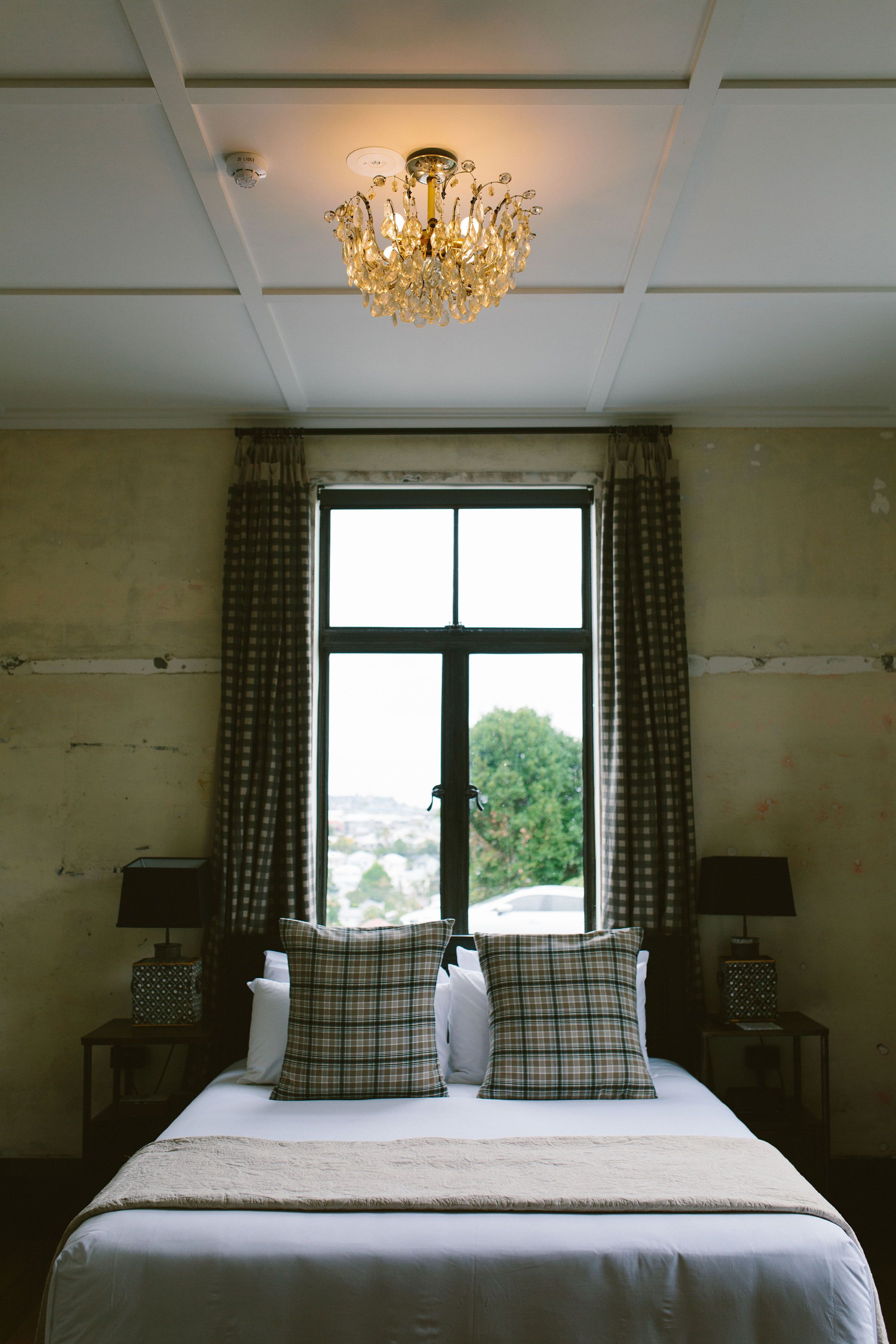 A bedroom with a large bed, two bedside tables, a chandelier, and a window with curtains.