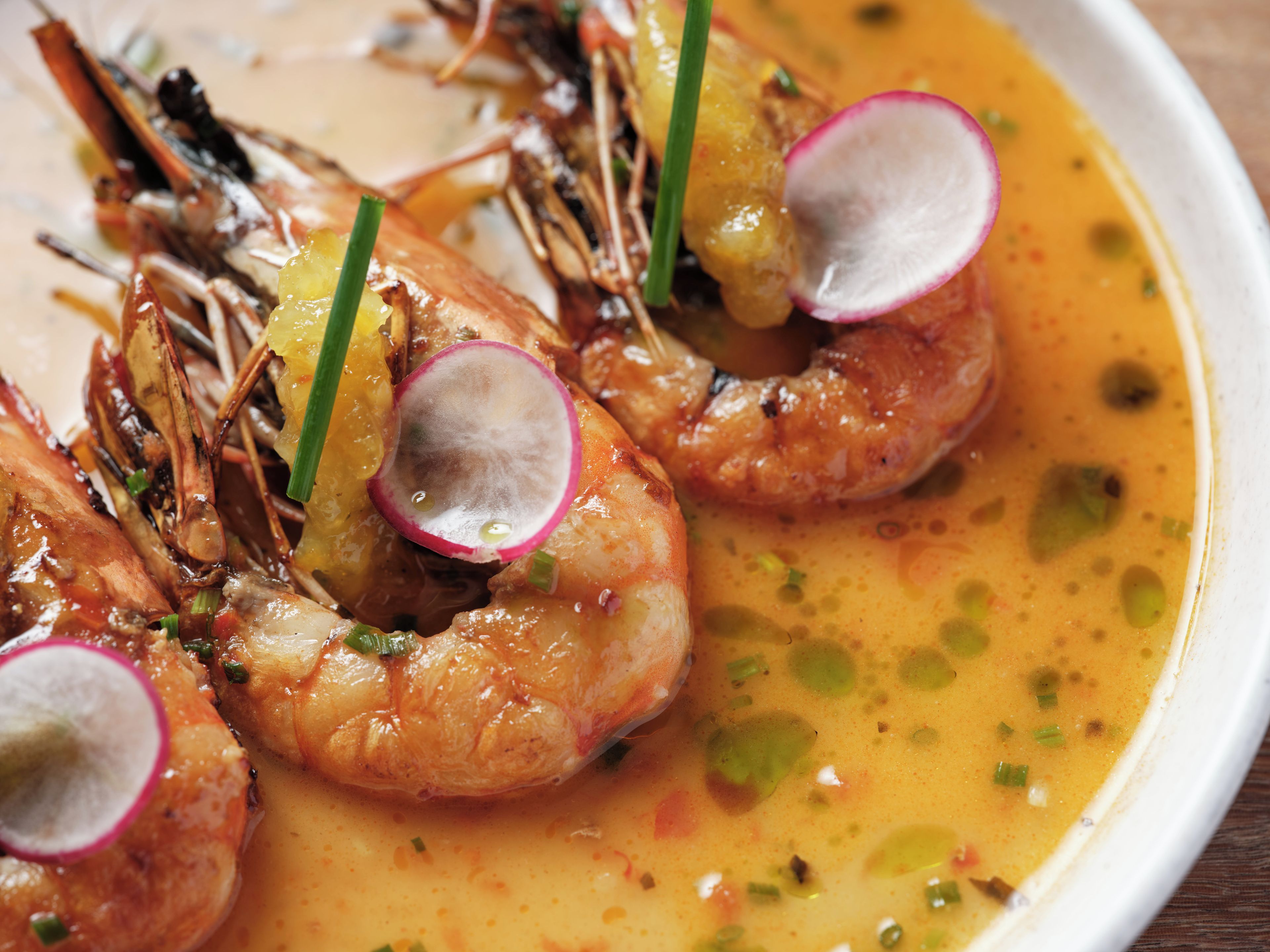 Une assiette de crevettes cuites, garnie de tranches de radis et d’oignons verts, servie dans un bouillon de couleur jaunâtre.
