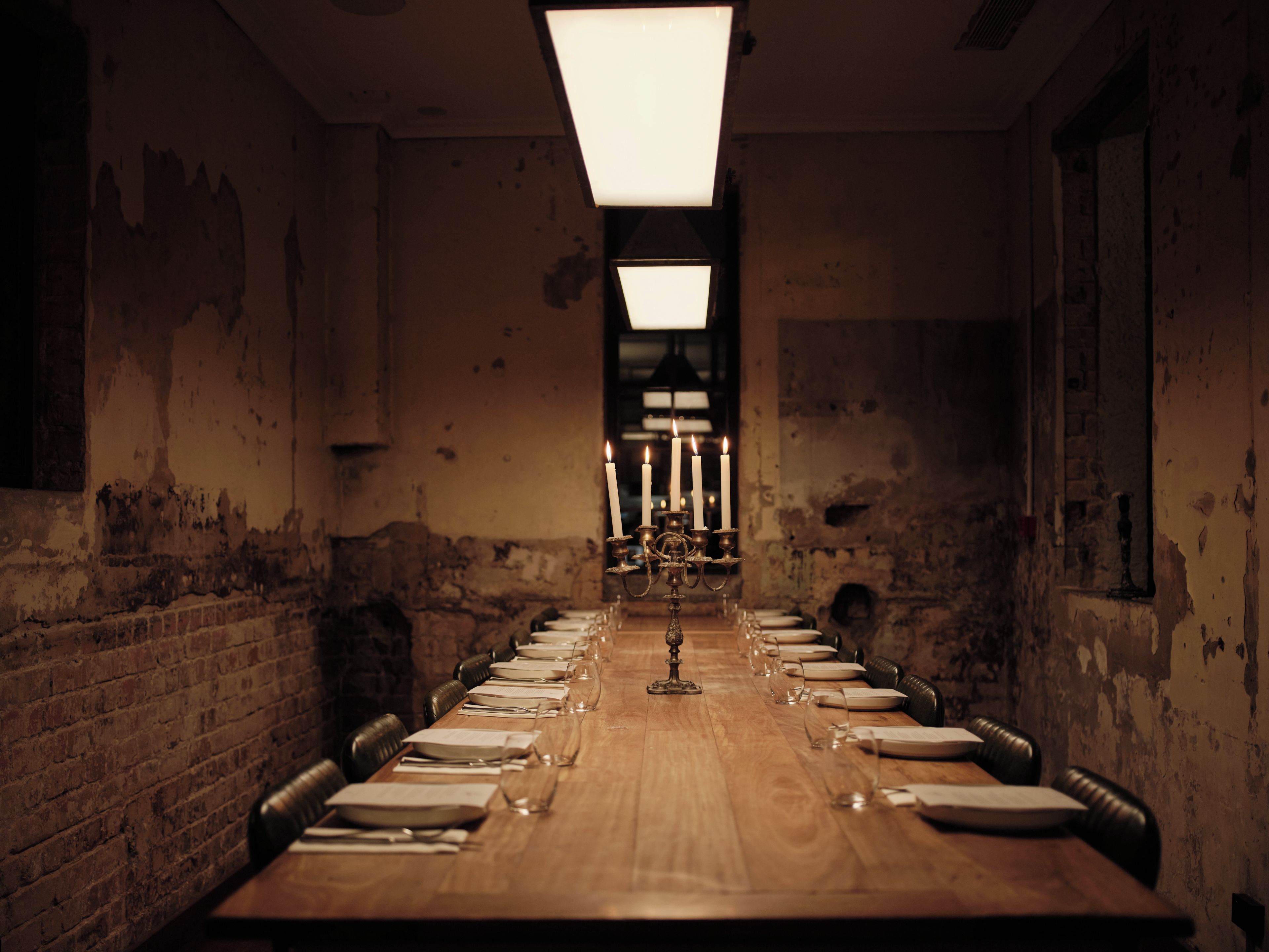 Une longue table en bois, dressée pour le dîner avec des bougies, des verres et des assiettes, dans une pièce aux murs de briques et au plafond haut.
