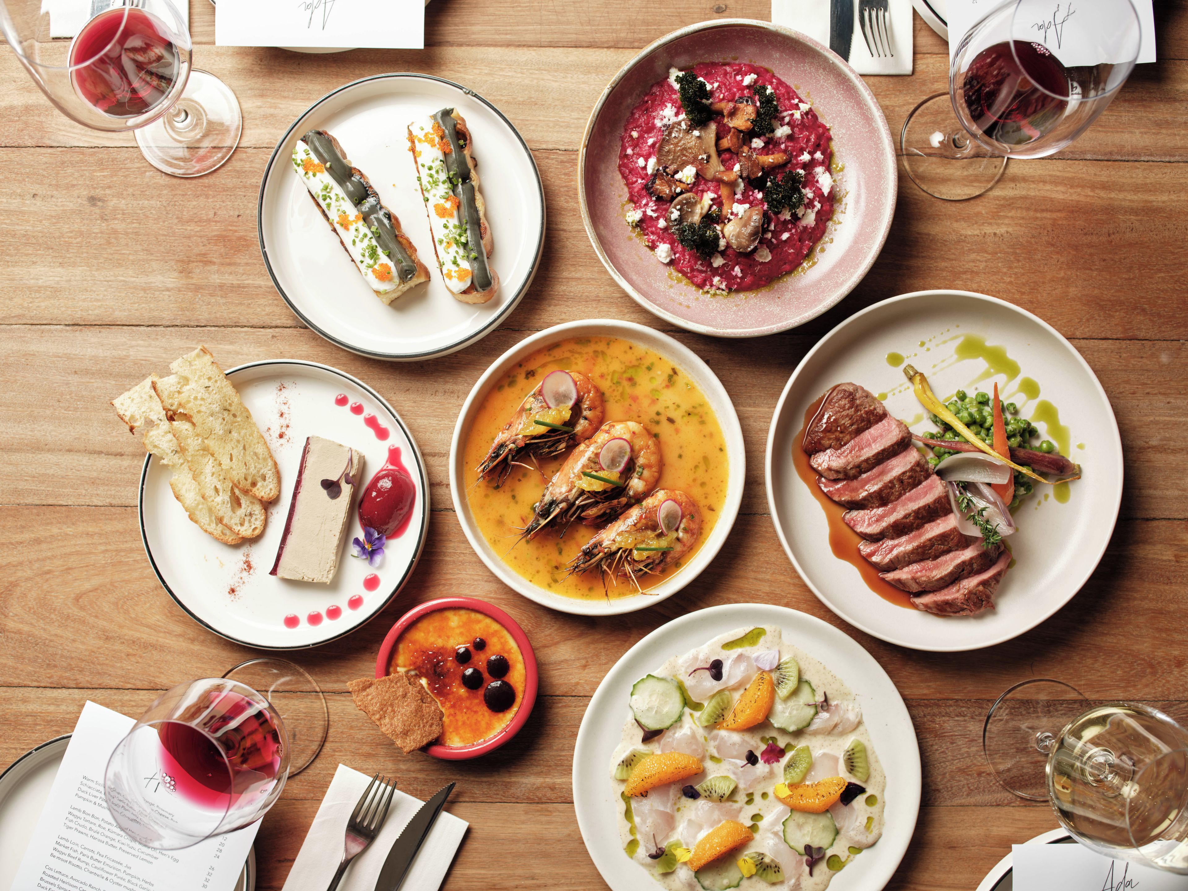 Variedad de platos sobre una mesa de madera, incluyendo mariscos, carne y verduras, con copas de vino y cubiertos.