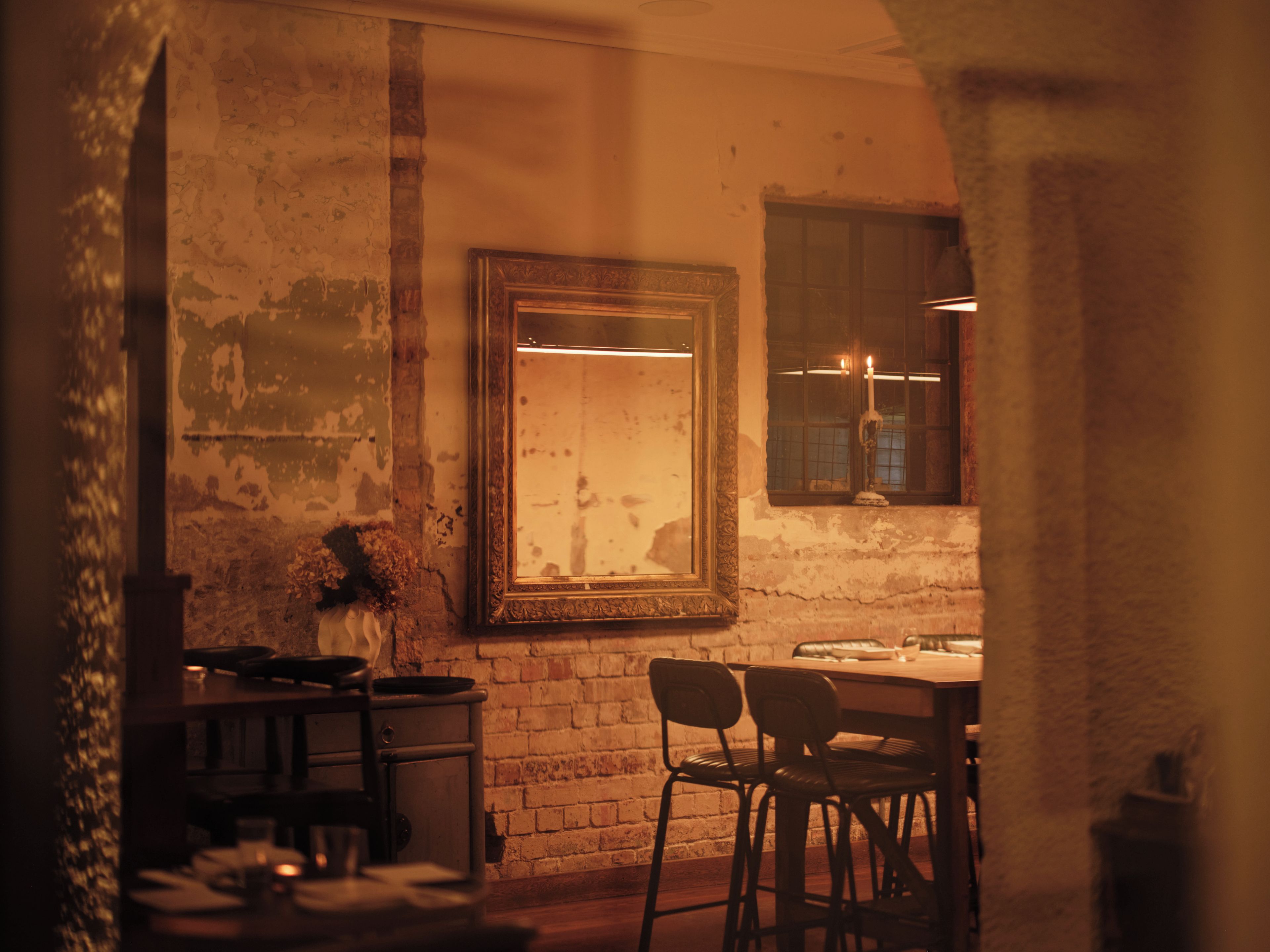 Una habitació poc il·luminada amb una paret de maó rústica, un mirall amb marc i una taula amb cadires.