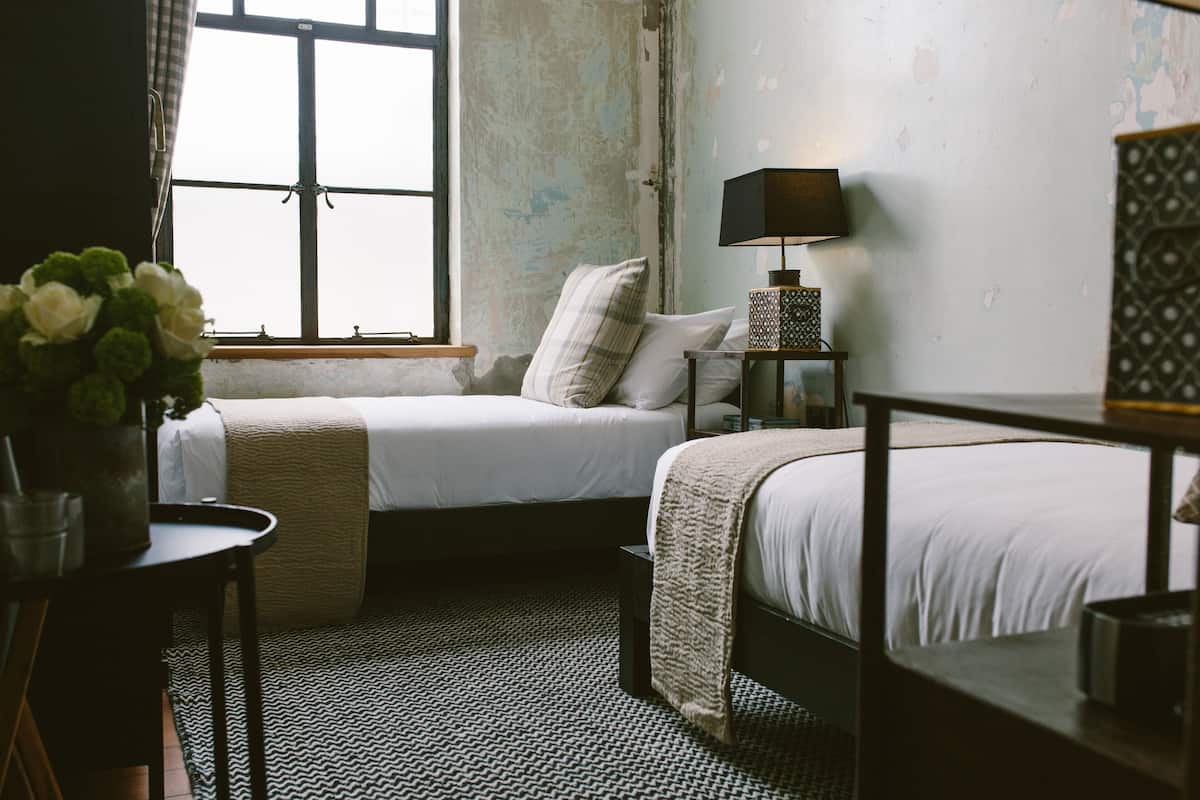 A room with two beds, a small table with a lamp, and a window with curtains.