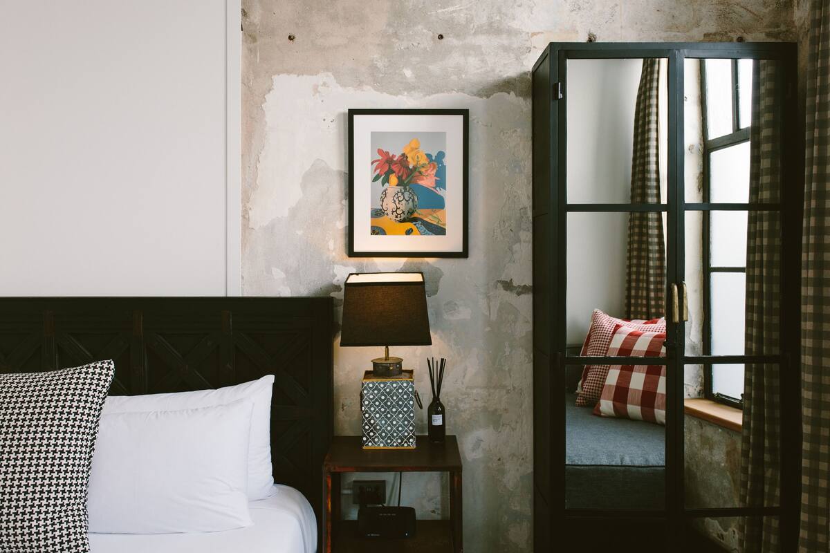 A bedroom with a bed, a bedside table with a lamp, a framed artwork, and a window with curtains.