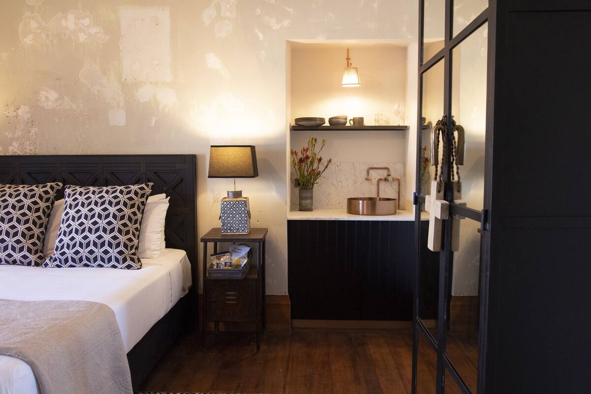 A bedroom with a bed, bedside table, lamp, and a small kitchenette area with a sink and countertop.