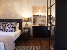 A bedroom with a bed, bedside table, lamp, and a small kitchenette area with a sink and countertop.