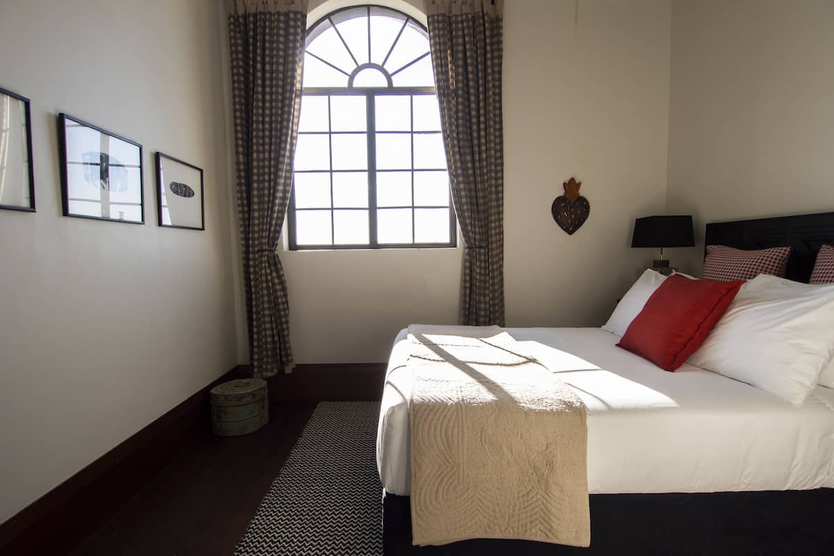 A bedroom with a large bed, a window with curtains, a wall-mounted lamp, and framed pictures.