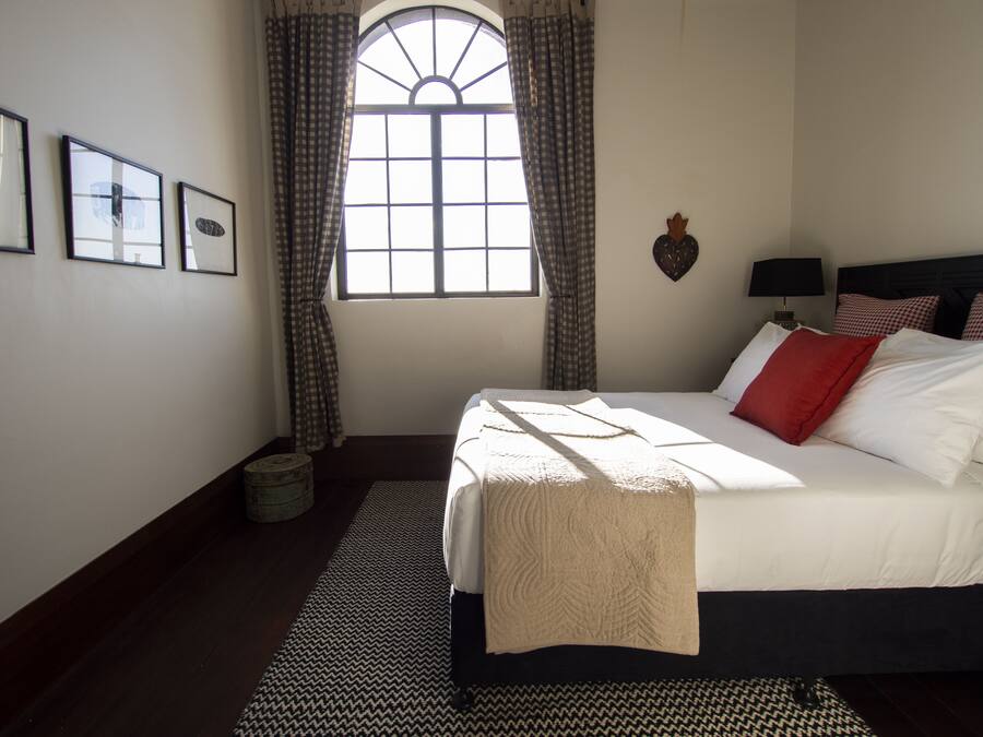A bedroom with a large bed, a window with curtains, a wall-mounted lamp, and framed pictures.