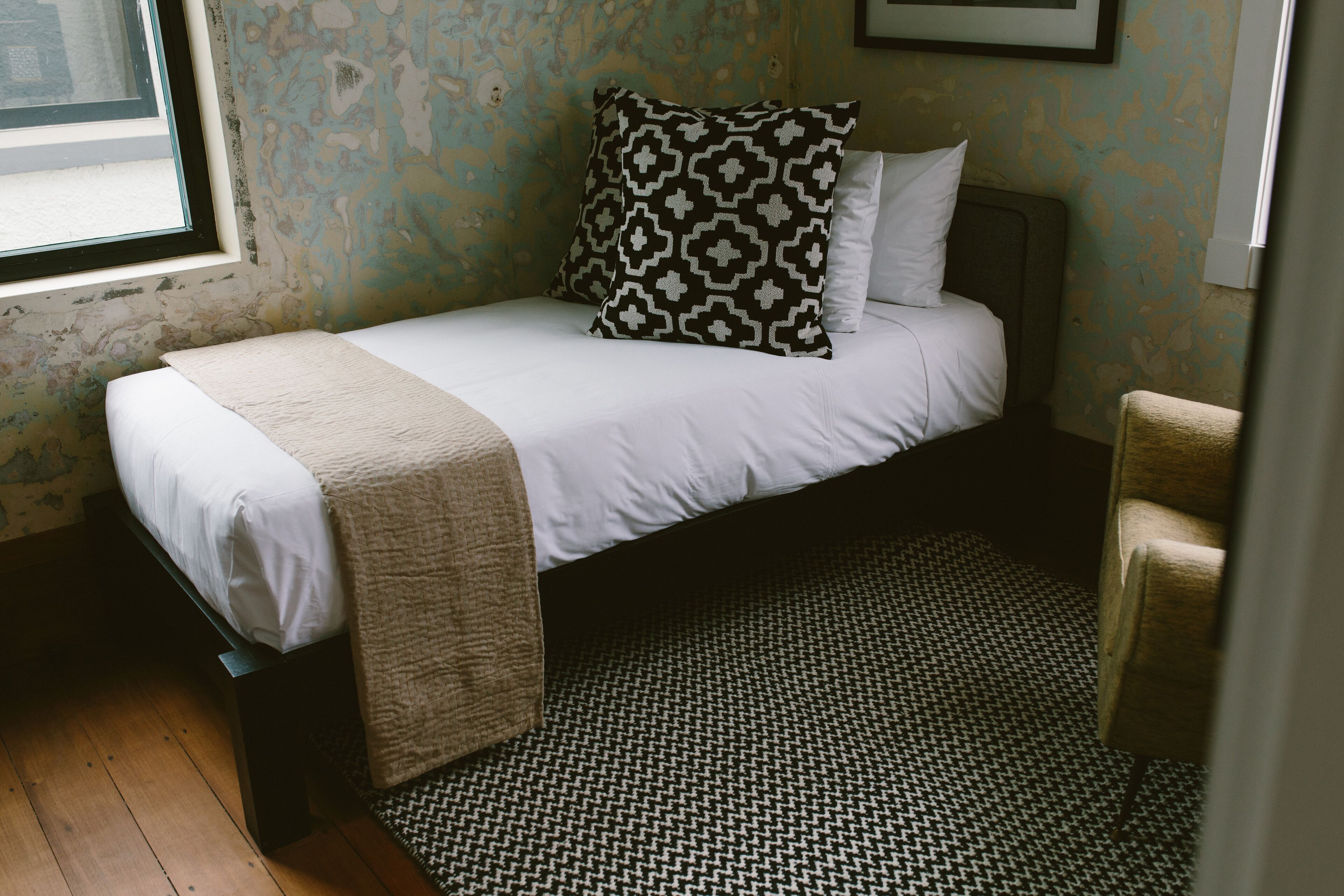 Een enkel bed met een gestreept kussen en een witte deken, een rug met een hondentandpatroon, en een stoel in een kamer met een raam en een muur met afbladderende verf.