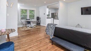 A modern, well-lit room with a wooden floor, a sofa, a small table, and a kitchenette in the background. - Cool City Cottage (MINS FROM AIRPORT/CITY) 2B 1B (Stone Mountain)