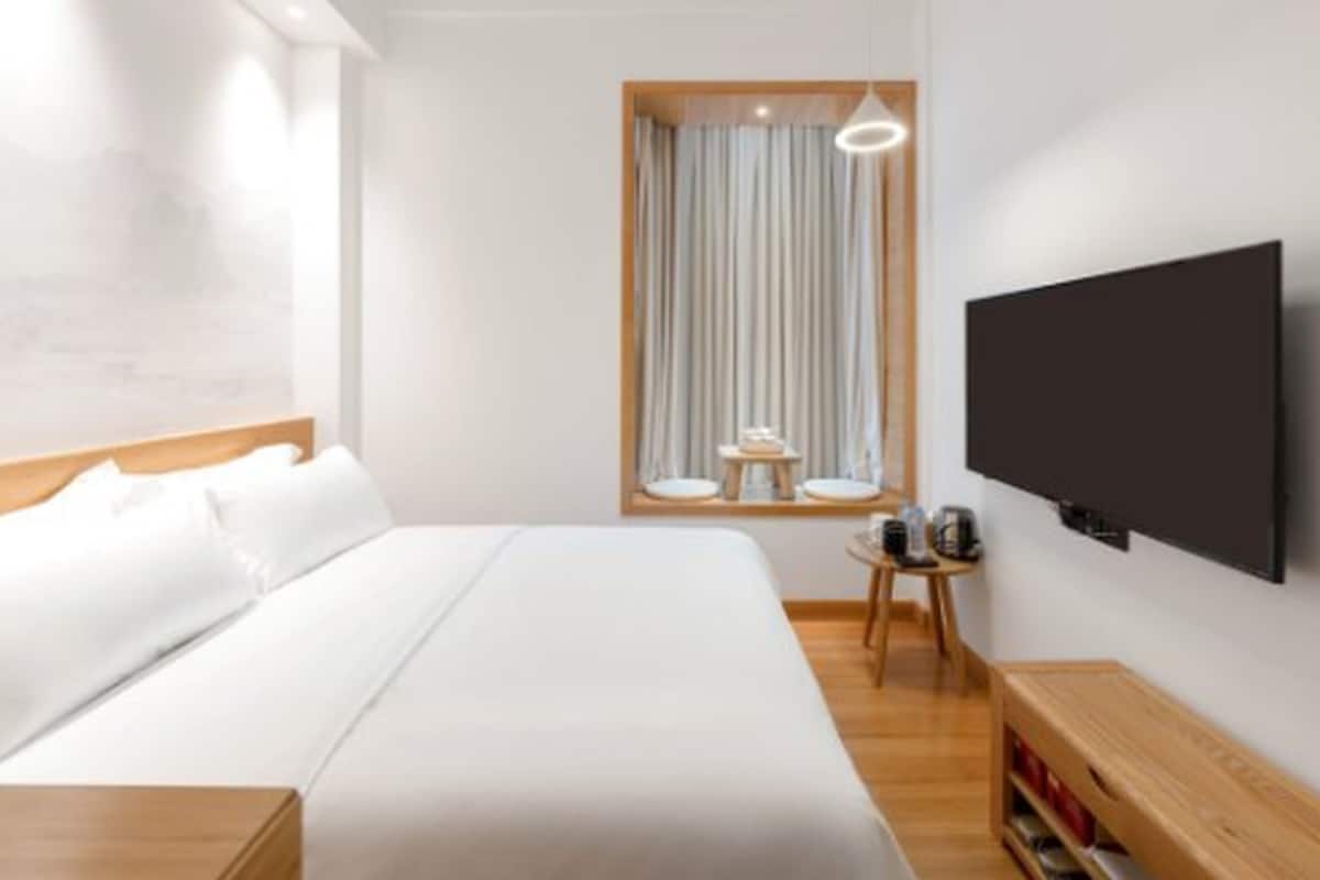 A modern hotel room with a large bed, a small bedside table with a lamp, a TV mounted on the wall, and a window with curtains.