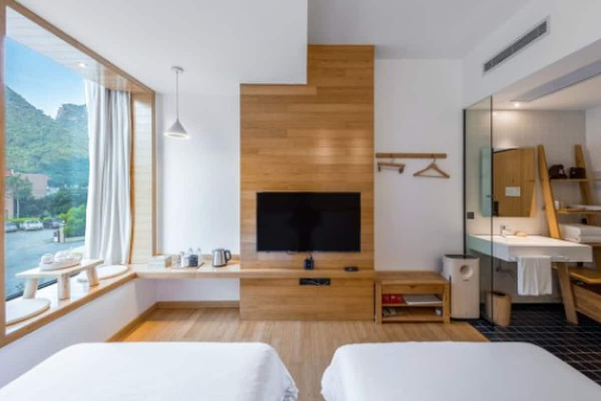 A modern hotel room with a large window, a flat-screen TV, wooden paneling, a small table with a kettle, and a bathroom with a mirror and towel rack.