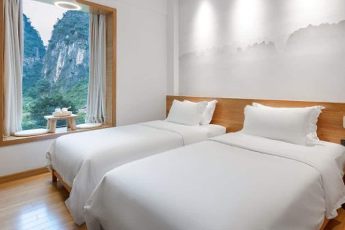 A hotel room with two beds, a wooden headboard, a nightstand, and a window with a view of a mountainous landscape.