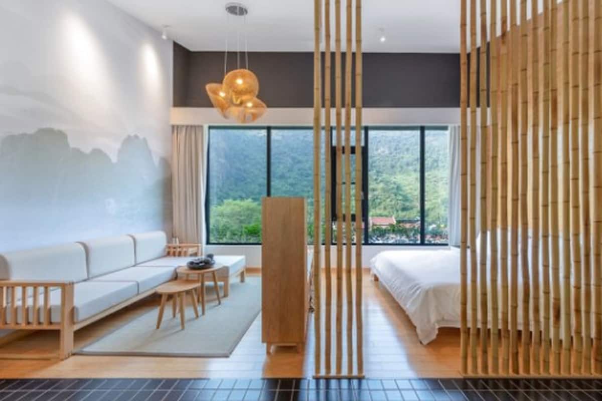 A modern hotel room with a sofa, a bed, a wooden partition, and a view of greenery outside.