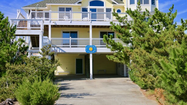 A two-story building with a balcony, a sign with a sun and palm tree, and a clear sky. - WH852- Stunning! Private Pool, Hot Tub, 4 BRs, Pool Table and Screened Porch! (Corolla)
