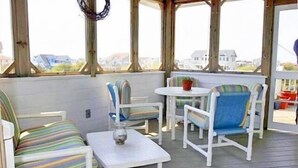 A covered outdoor seating area with white furniture and a small table, overlooking a residential neighborhood. - WH852- Stunning! Private Pool, Hot Tub, 4 BRs, Pool Table and Screened Porch! (Corolla)