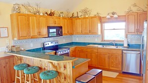 A kitchen with wooden cabinets, a central island with stools, a microwave, stove, sink, and dishwasher. - WH852- Stunning! Private Pool, Hot Tub, 4 BRs, Pool Table and Screened Porch! (Corolla)