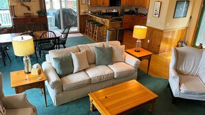A living room with a sofa, a coffee table, a dining area with chairs, a kitchen with wooden cabinets, and a sliding glass door leading to an outdoor area. - WH852- Stunning! Private Pool, Hot Tub, 4 BRs, Pool Table and Screened Porch! (Corolla)