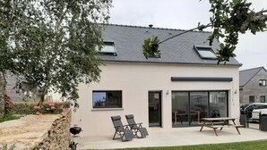 Ein modernes Haus mit einer Terrasse, zwei Liegestühlen, einem Holztisch und einem Grill.