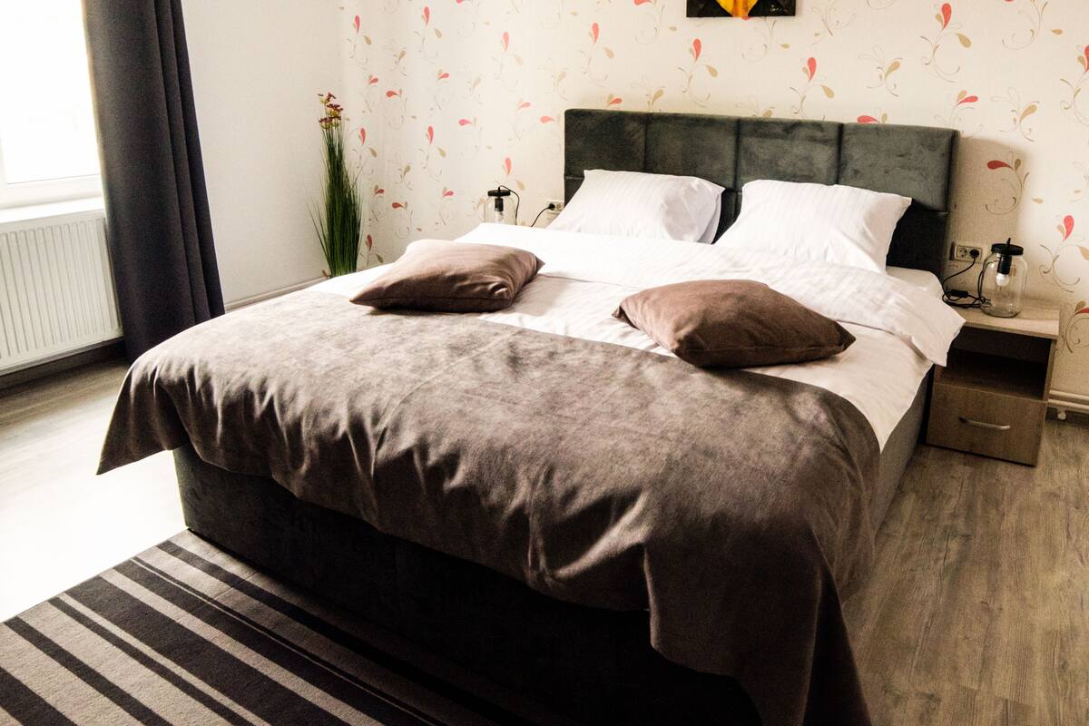 A neatly made bed with a dark brown bedspread and two pillows, a nightstand with a lamp, a wall with a floral pattern, and a window with a curtain.