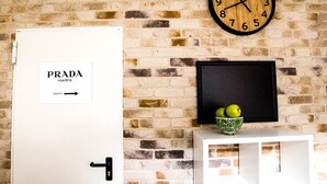 A room with a brick wall, a clock, a door labeled "PRADA MARFA," and a small white console table with a bowl of green apples.