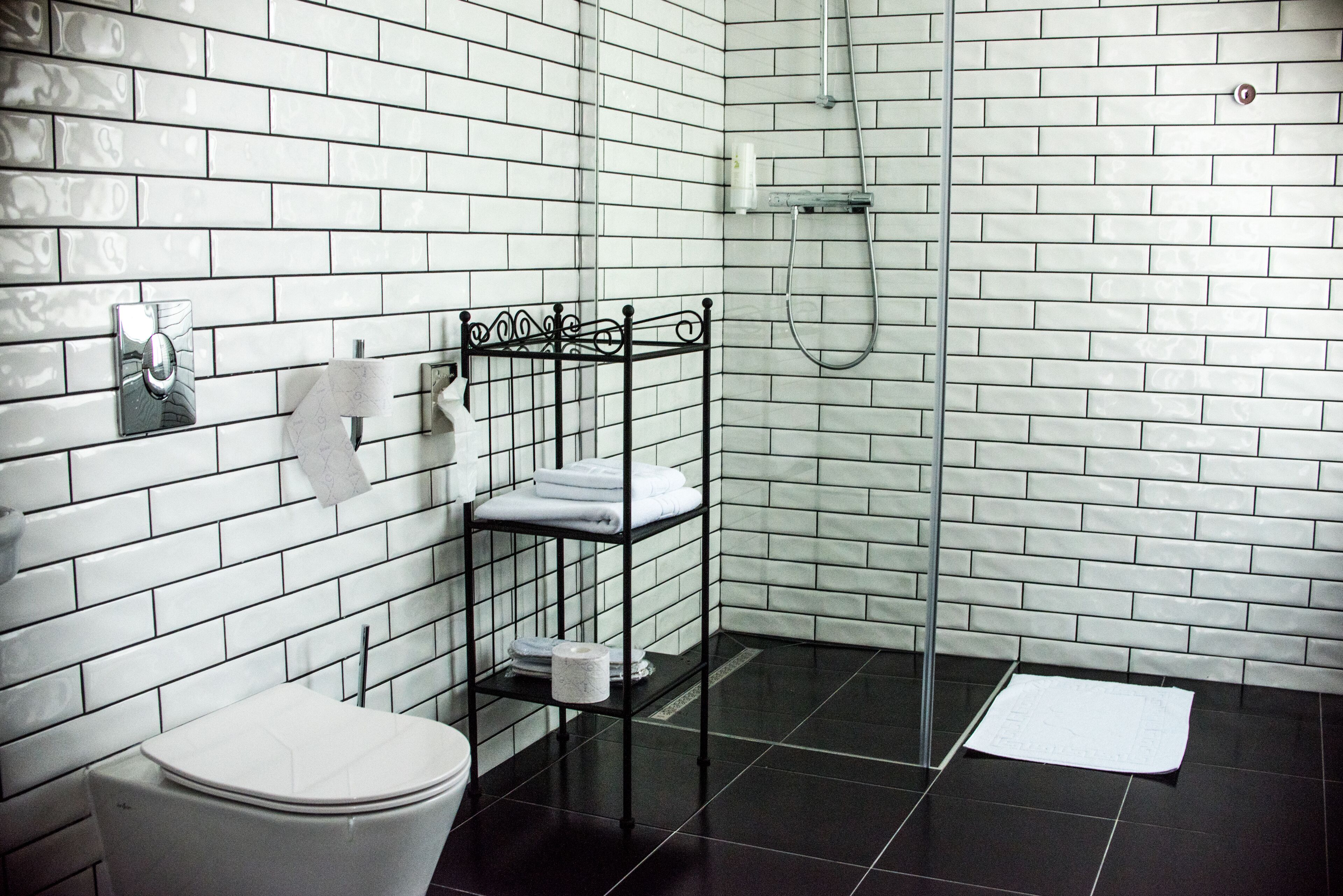Ein Badezimmer mit weißen Fliesen, schwarzem Boden, Toilette, Ablage mit Handtüchern und einer Duschkabine.