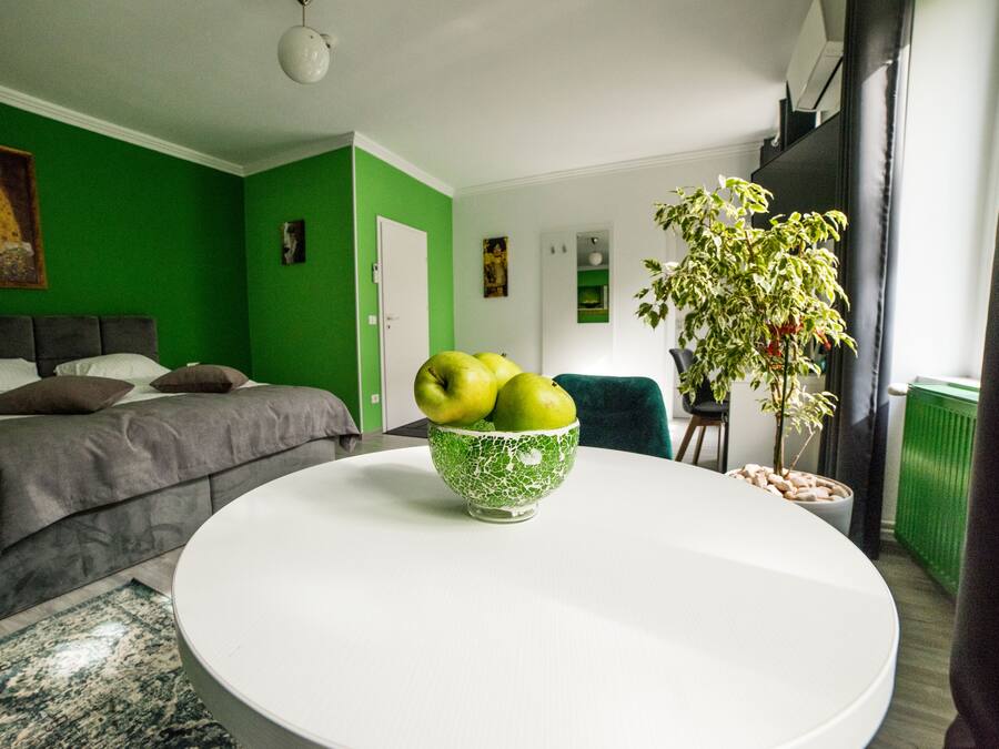 Un dormitorio moderno con una pared de acento verde, una cama con ropa de cama gris, una mesita redonda blanca con un bol verde de manzanas y una planta en maceta.
