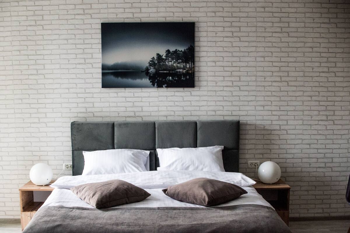 A bedroom with a white brick wall, a grey upholstered headboard, a large bed with white and brown bedding, and two bedside tables with white spherical lamps.