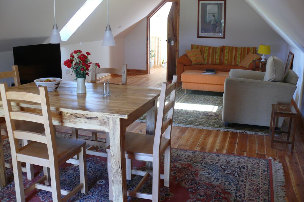 A cozy living room with a wooden dining table, chairs, a sofa, and a patterned rug.