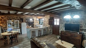 A rustic kitchen with stone walls, wooden beams, a central island with stools, a dining table with chairs, and a TV on a wooden cabinet. - A Casa dos Avós - House with swimming pool, barbecue in the middle of nature in A Coruña. (Oza de los Ríos)