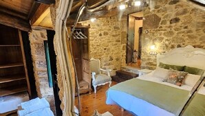 A rustic bedroom with a stone wall, wooden floor, a bed with green bedding, a white chair, and a mirror reflecting the room. - A Casa dos Avós - House with swimming pool, barbecue in the middle of nature in A Coruña. (Oza de los Ríos)