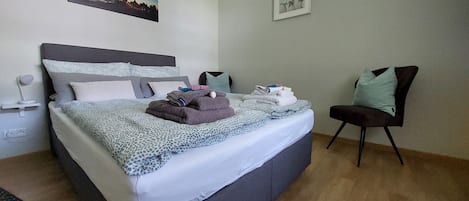 Una cama bien hecha con una colcha estampada, cabecera y almohadas. Hay una silla con cojín y una mesita de noche con una lámpara.