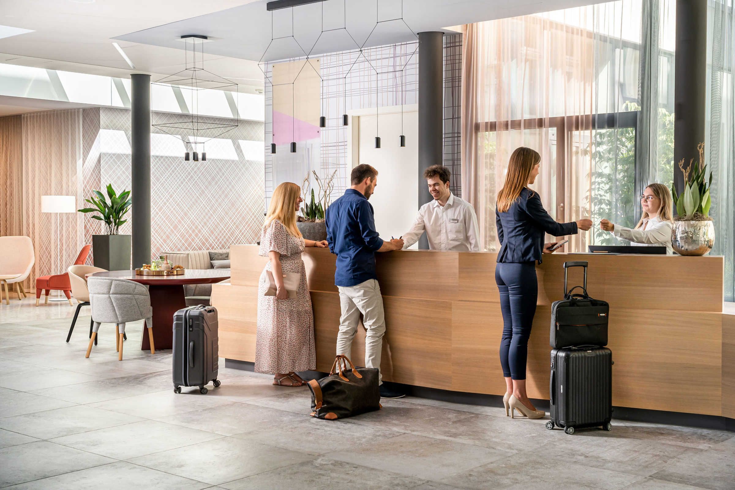Una moderna area reception hotel con una scrivania in legno, un angolo salotto con divano e poltrone, e un'ampia finestra che offre luce naturale.