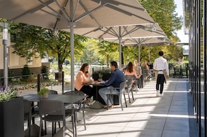 An outdoor café with tables and chairs under umbrellas, surrounded by trees and a pathway.