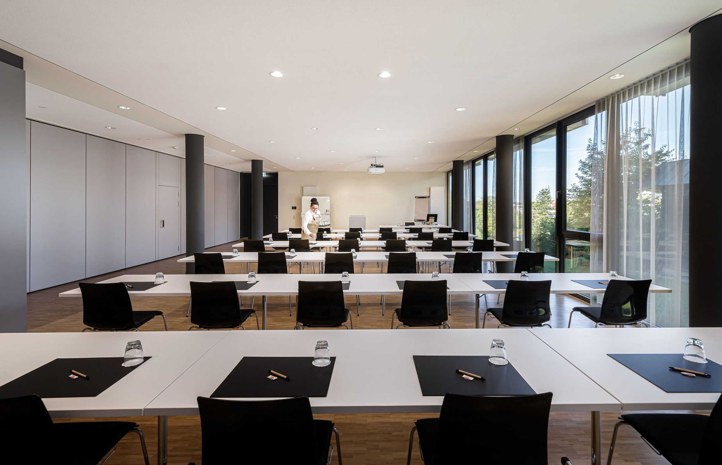 A conference room with multiple tables and chairs, large windows, and a projector screen.