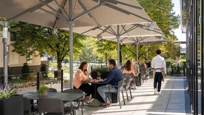Un café al aire libre con mesas y sillas bajo sombrillas, rodeado de vegetación y edificios.