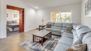 A spacious living room with a large gray sectional sofa, a wooden coffee table, and a patterned rug. There is a door leading to a bedroom with a bed and a small table. - Outdoor Oasis: Coachella Valley Group Getaway! (Coachella)